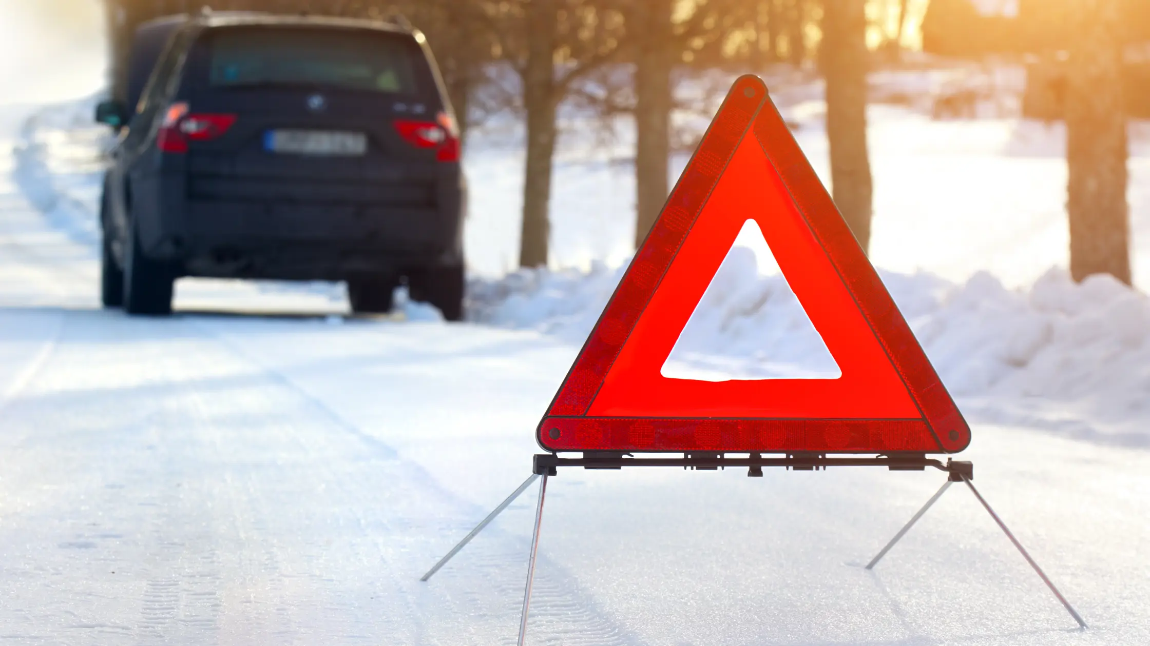 Orange varningstriangel ståendes på snötäckt väg med en mörk bil i bakgrunden. Guldigt solljus skiner mot kameran.