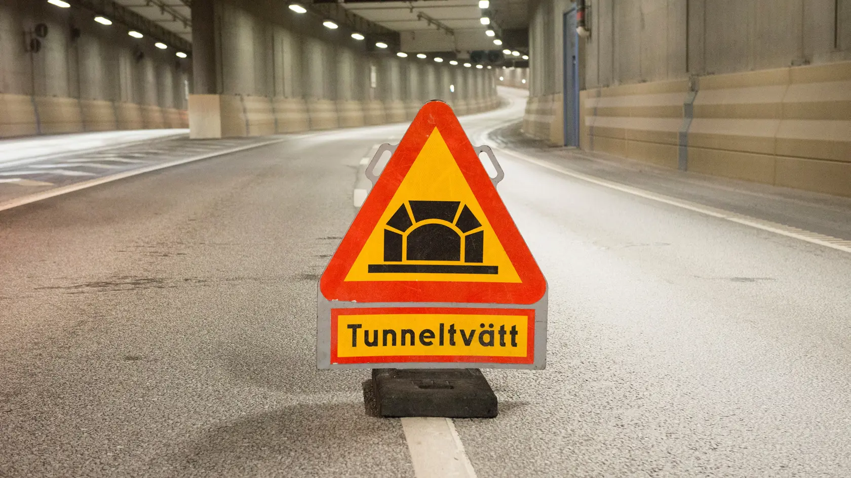 Vägskylt varning för tunneltvätt. Står på vägbanan i en tunnel som mynnar ut i två rör i bakgrunden.