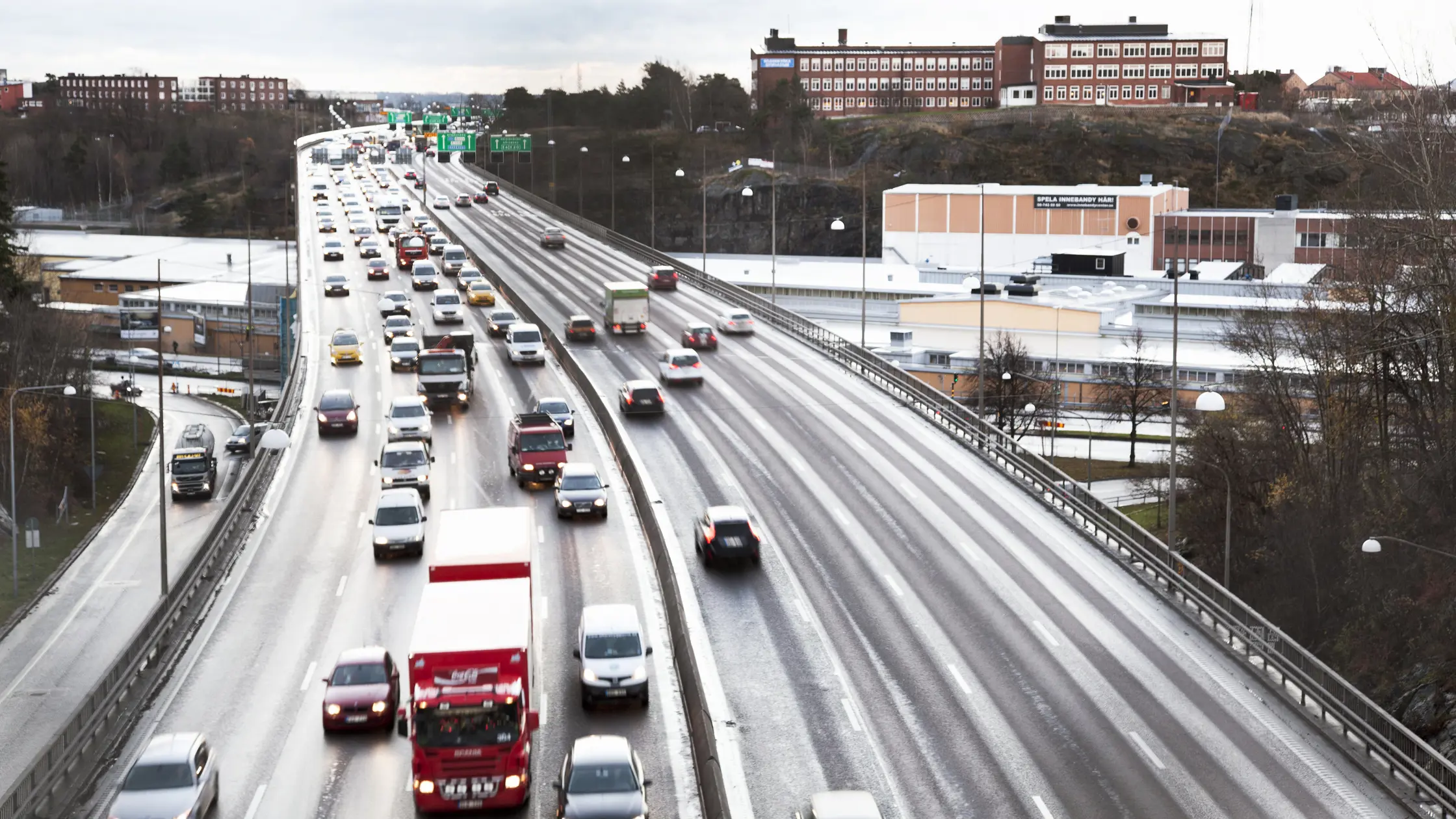 Bild tagen på del av Essingeleden ovanifrån. Tung trafik delar utrymme med personbilar. Byggnader syns i bakgrunden.