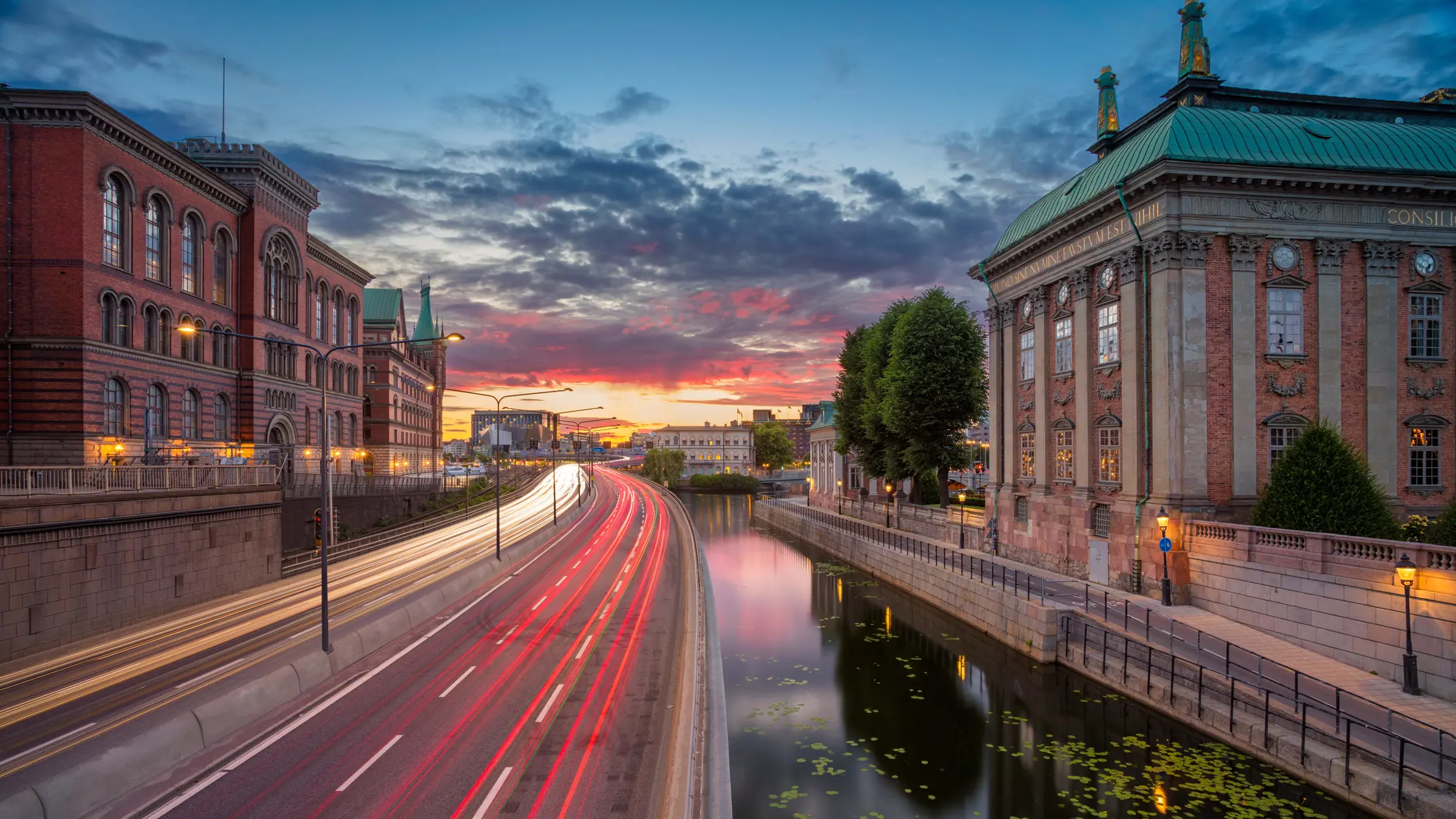 Stockholm, sommar, solnedgång, trafik som flyter som röda och vita sträck. Vatten på höger sida och gamla hus som tornar upp sig på vardera sida.