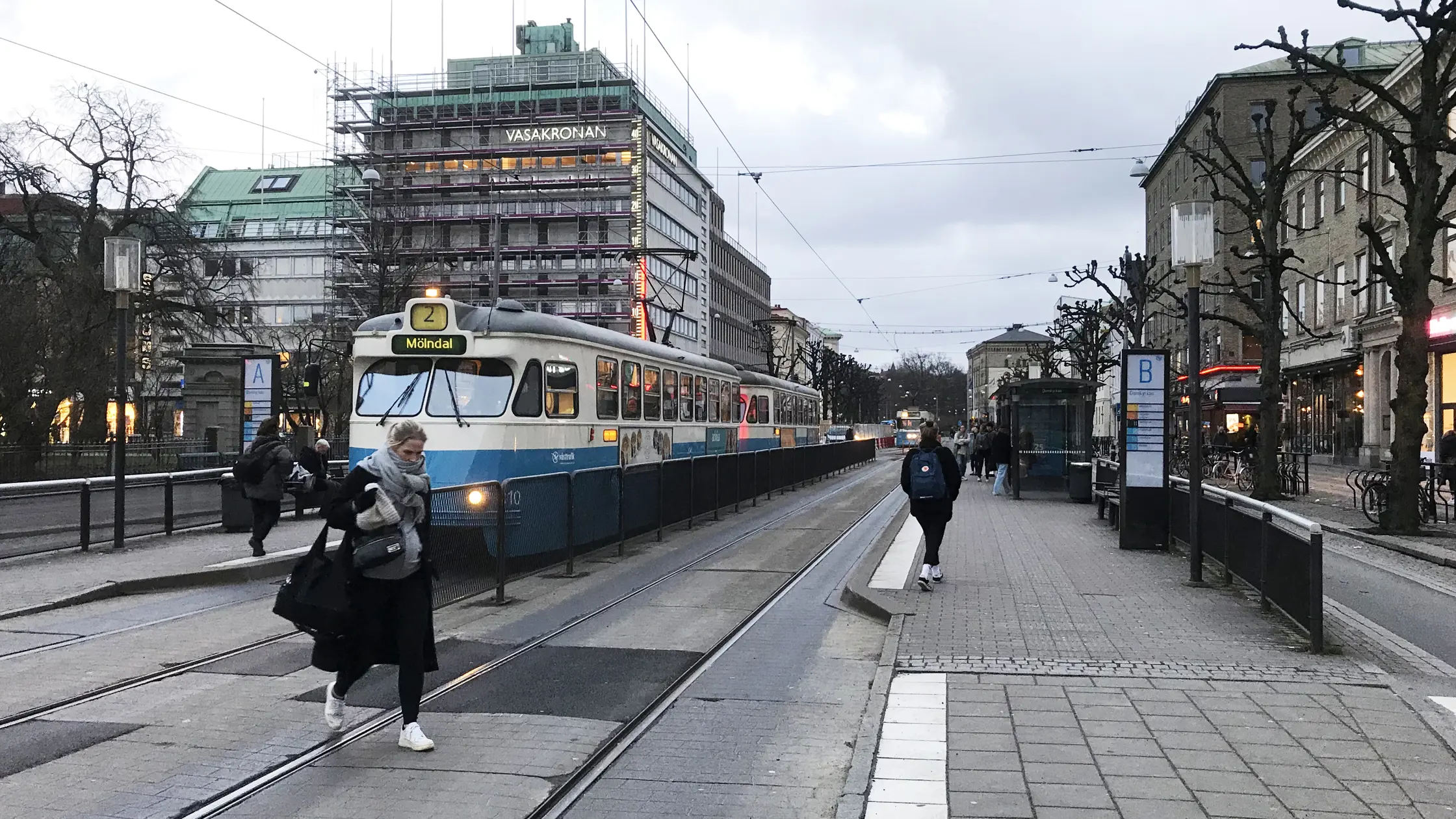 Personer syns gå över övergångsstället och längs perrongen vid hållplats Domkyrkan. Spårvagnslinje 2 står still vid hållplatsen