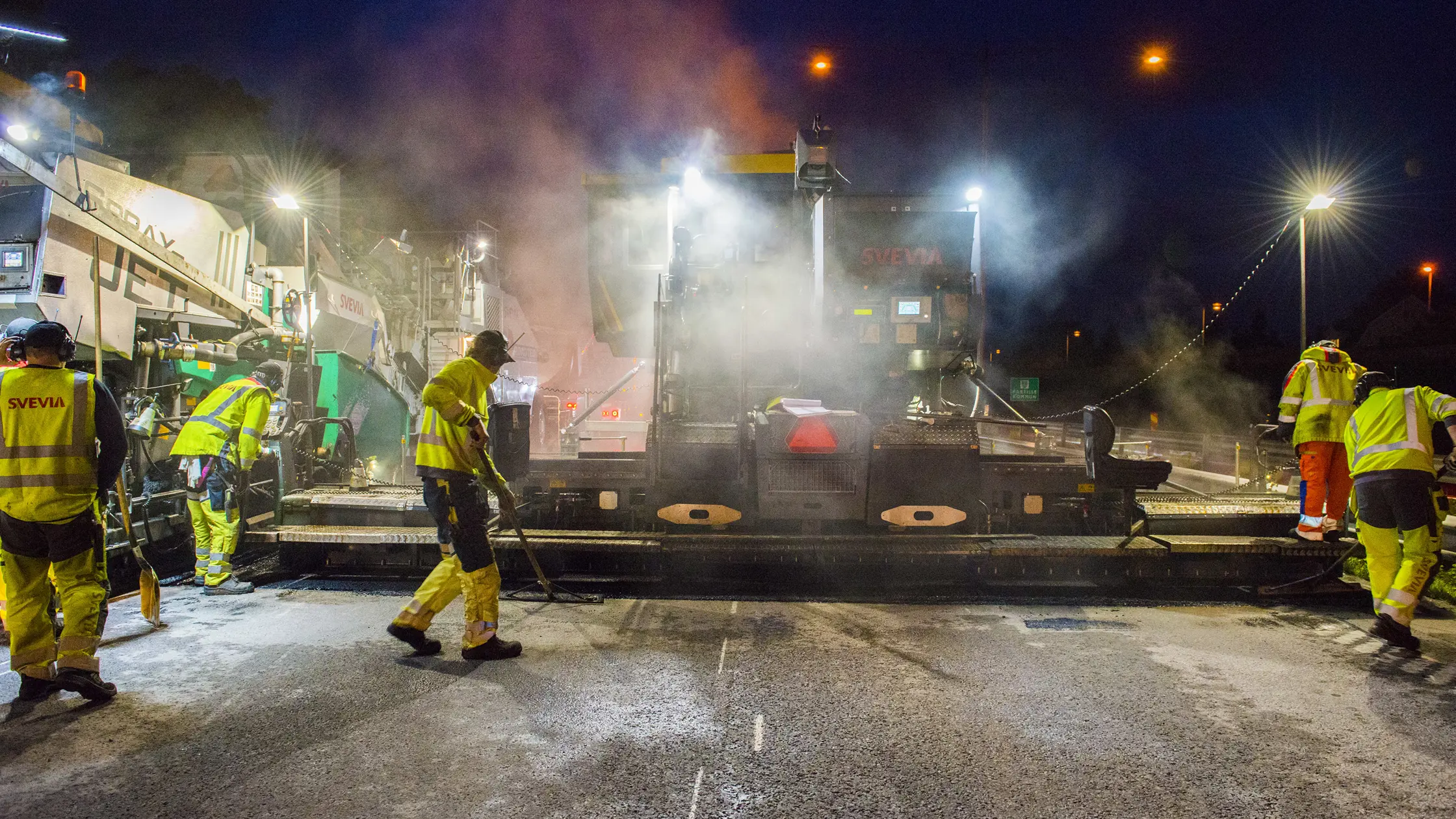 Asfalteringsarbete pågår nattetid där ett flertal vägarbetare deltar. 