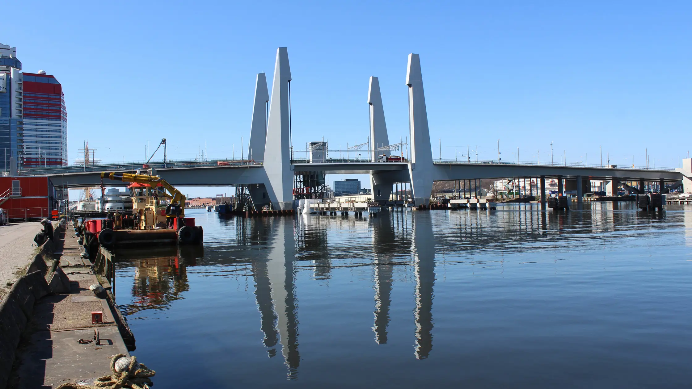 Nästan hela Hisingsbron sedd från kajen på fastlandet vid Holmen, en solig dag där pylonerna speglas i vattnet. 
