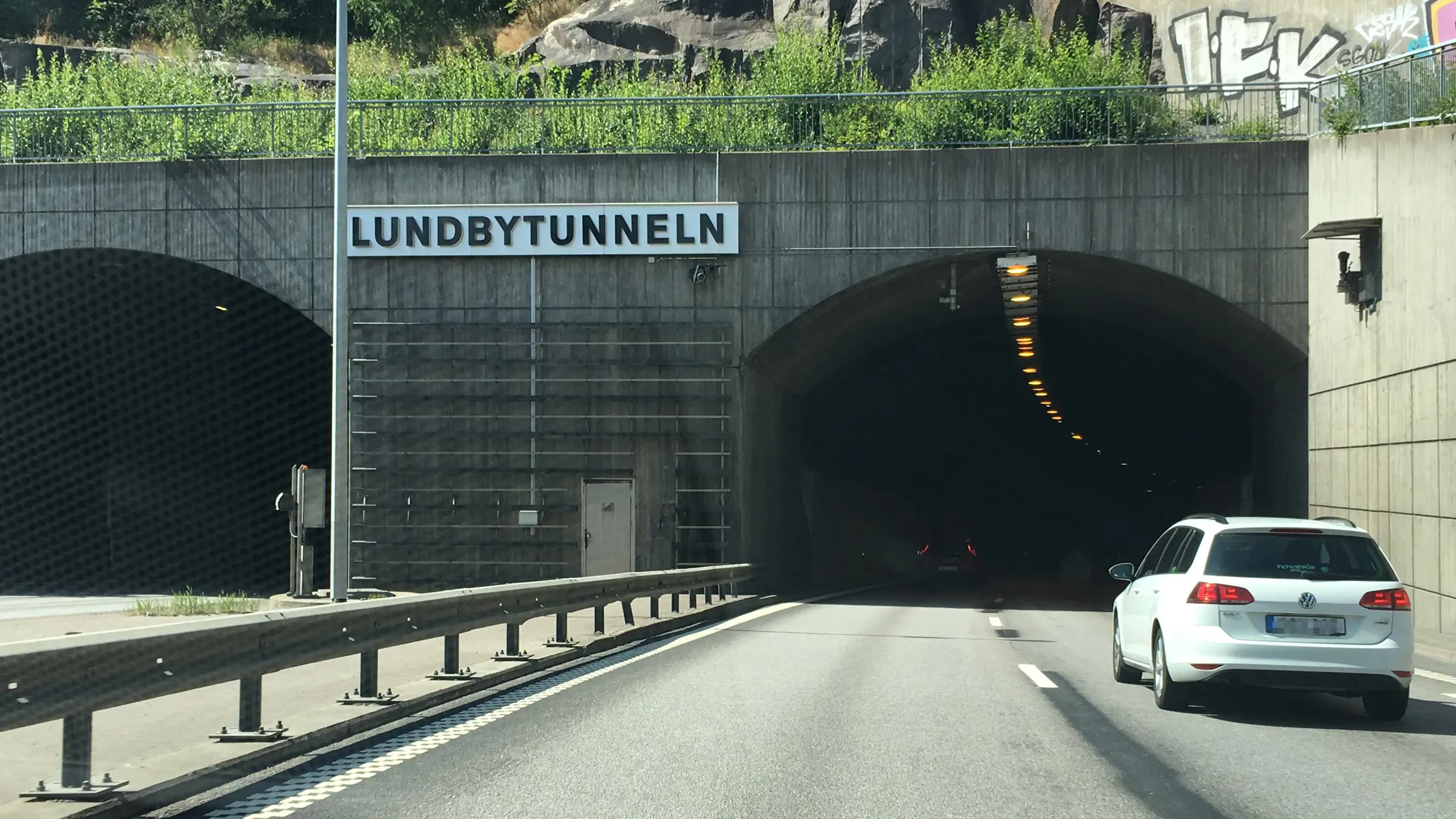 Vit bil kör in i Lundbytunneln. I bakgrunden syns skylten Lundbytunneln och tunnelmynningen. 