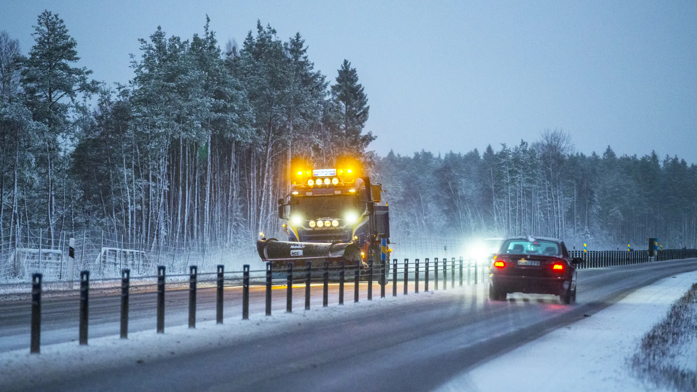 En plogbil är ute och kör på en motorväg med vajeräcke en mötande rödbil passerar.